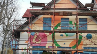 Przy budynku stoi rusztowanie. Na ścianie widać nieskończony kolorowy mural