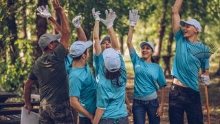 Grupa osób w błękitnych koszulkach wydaje okrzyk radości. Znajdują się w ogrodzie. Wokół nich są narzędzia ogrodnicze. Realizują wolontariat pracowniczy.