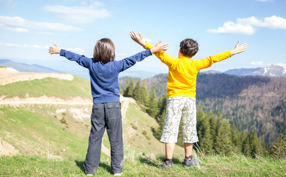 Dwójka dzieci stoi na wzniesieniu z rękoma wzniesionymi ku górze. Przed nimi malowniczy, górski krajobraz