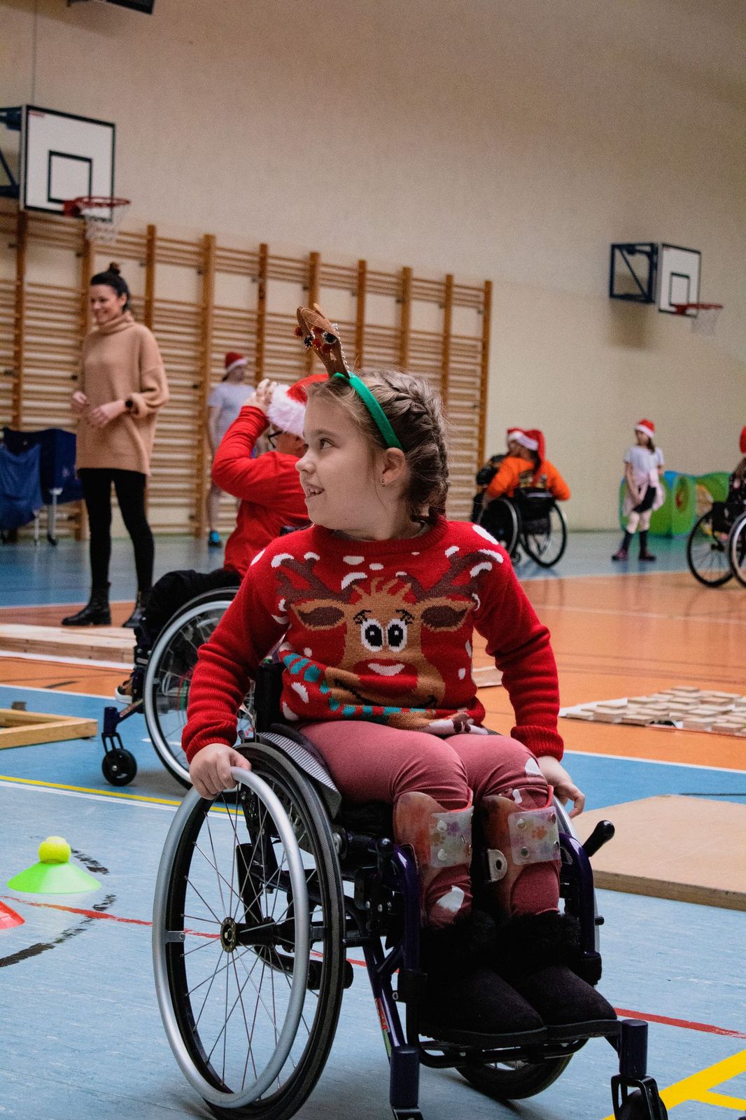 Dziewczynka w świątecznym swetrze i rogach renifera odwraca się do kogoś kto stoi po jej prawej stronie. Dziewczynka porusza się na wózku.
