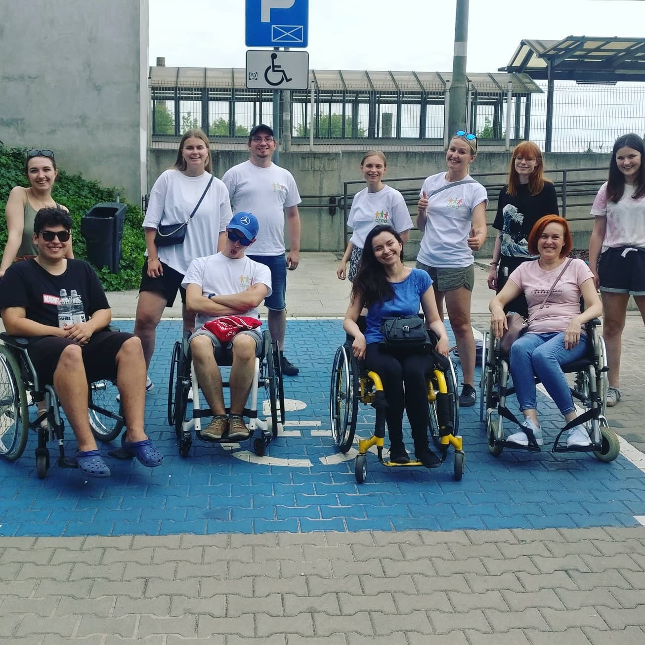 Grupa osób, w tym część na wózkach stoi na miejscu parkingowym dla osoby z niepełnosprawnością. Uśmiechają się.