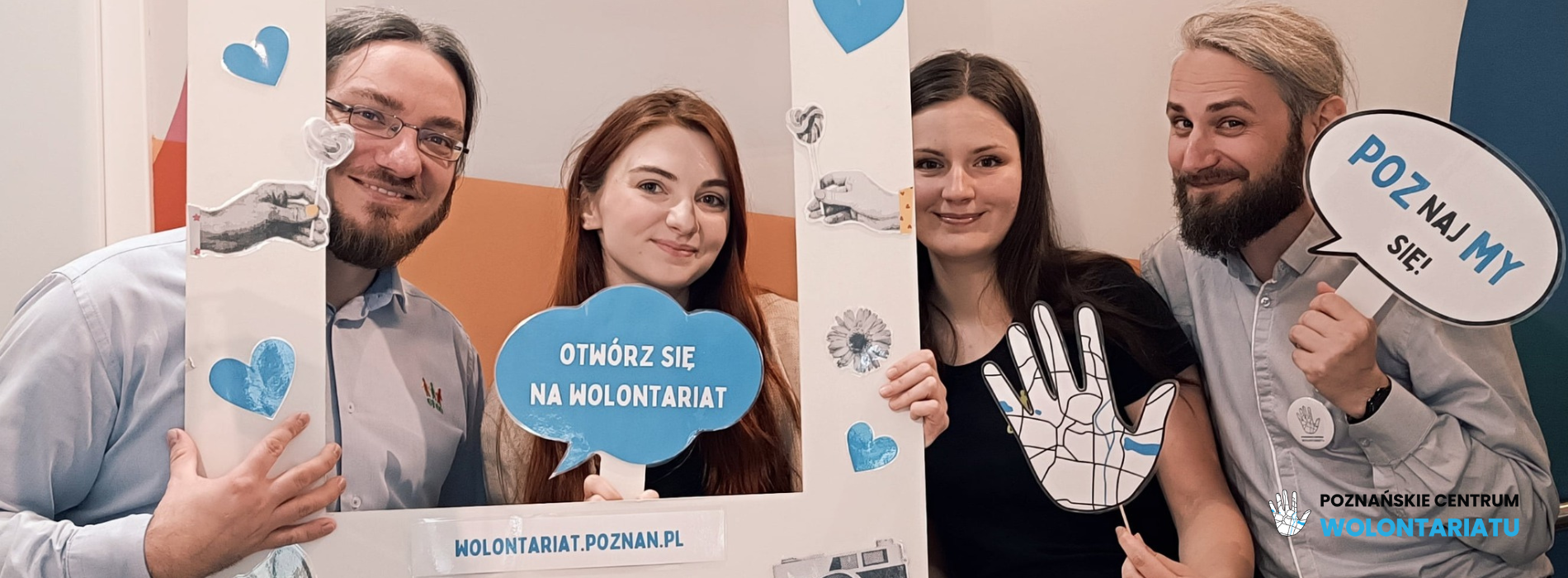 cztery uśmiechnięte osoby. Dwóch mężczyzn i dwie kobiety. Trzymają w dłoniach banery z napisem Poznańskie Centrum Wolontariatu. Pozują w ramce do zdjęć.
