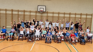 Grupa osób ustawiona do zdjęcia na sali gimnastycznej. Z przodu ustawione osoby na wózkach. Wszyscy machają do kamery.