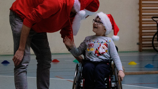 Młody mężczyzna w czapce Mikołaja pochyla się nad uśmiechniętym chłopcem, który porusza się na wozku.