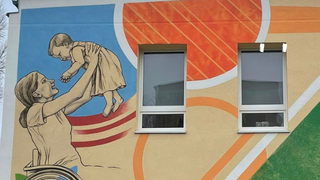 Na murali widzimy matkę, które z zachwytem unosi swoje małe dziecko ku górze. Kobieta porusza się na wózku