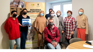 Grupa osób pozuje przed banerem z napisem Akademia Wolontariatu. Stoją w salce szkoleniowej i uśmiechają się. Jeden z mężczyzn jest na wózku.