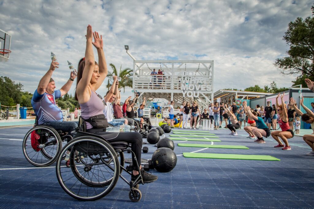 Grupa osób ćwicząca crossfit. Część osób jest na wózku, część chodząca. Stoją na przeciwko siebie.