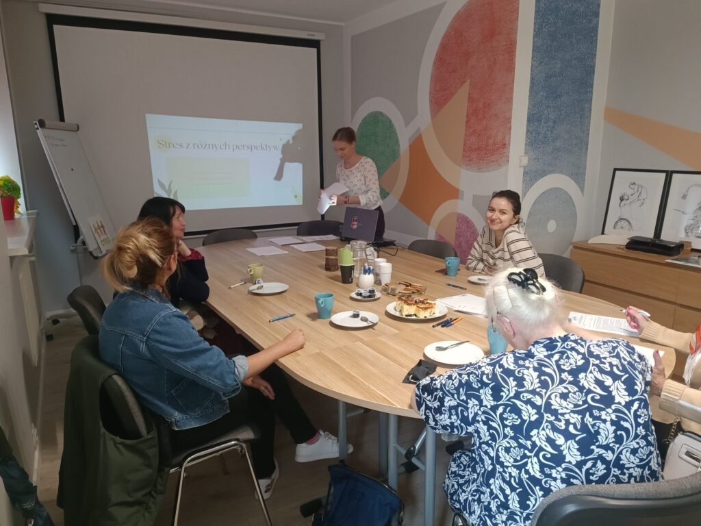 Grupa osób siedząca przy stole. Prelegentka warsztatów stoi. Za nią znajduję się tablica na której jest wyświetlany slajd: stres z różnych perspektyw