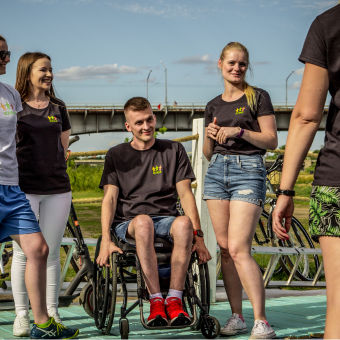 Trzy kobiety i jeden mężczyzna poruszający się na wózku śmieją się i rozmawiają. Ubrani są w czarne koszulki z logo CREO.