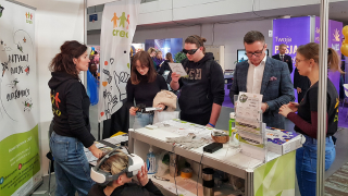 Przed stolikiem stoją 3 osoby, młoda dziewczyna i chłopak przymierzają symulatory wzroku. Obok mężczyzna rozmawia z Michaliną. Za ladą Agata i Natalia patrząca w okulary wirtualnej rzeczywistości.