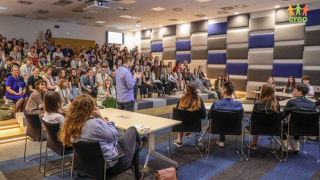 Sala konferencyjna. Po środku siedzą uczniowie którzy prowadzą debatę oxfordzką. Na środku stoi przygotowujący ich do debaty trener, który przedstawia uczniów. na przeciwko siedzą widzowie, w tym uczniowie i nauczyciele.