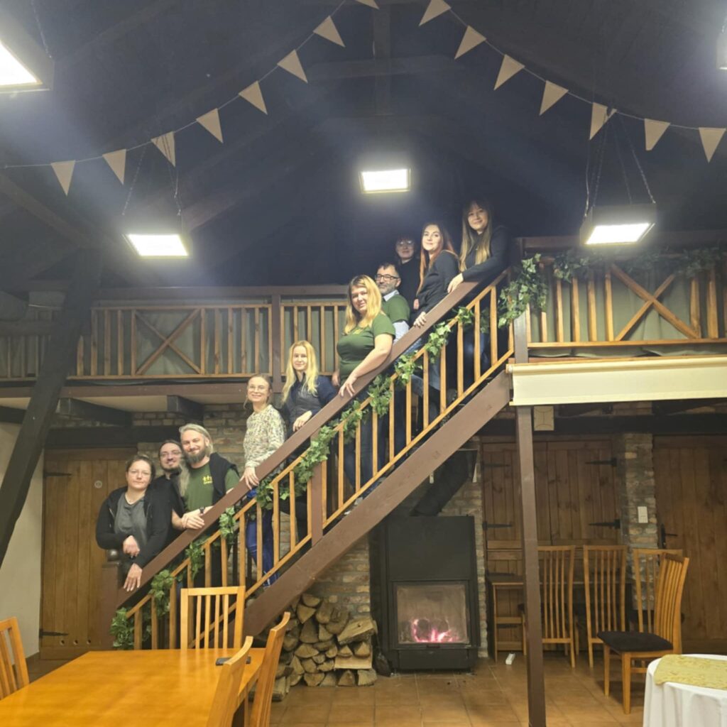 Grupa osób pozuje na drewnianych schodach w przytulnym, rustykalnym wnętrzu z kominkiem. Uśmiechnięci uczestnicy stoją w rzędzie, tworząc ciepłą, integracyjną atmosferę.