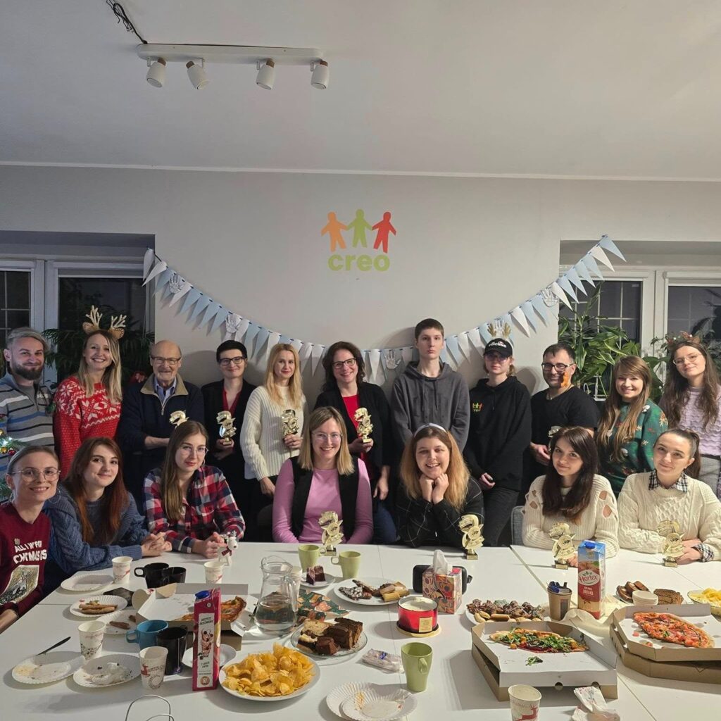 Na zdjęciu grupa ludzi pozuje do wspólnego zdjęcia przy świątecznie udekorowanym stole, pełnym jedzenia i napojów. W tle widać logo "Creo".