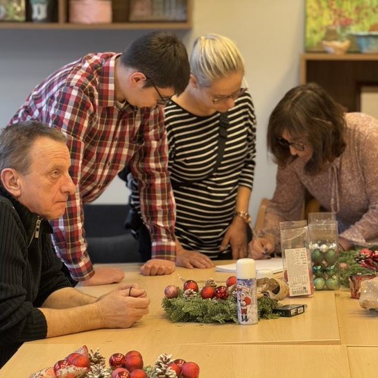 Grupa dorosłych osób pracuje wspólnie przy stole, przygotowując świąteczne dekoracje z bombkami i wieńcami. Jedna osoba siedzi zamyślona, a pozostałe trzy są zaangażowane w tworzenie ozdób lub notowanie.