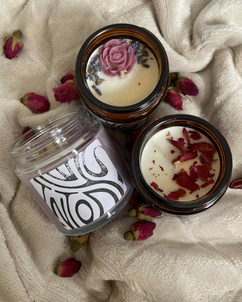 Trzy ozdobne świece w szklanych słoiczkach, ozdobione suszonymi kwiatami i ziołami, leżą na miękkim, kremowym kocu w otoczeniu suszonych pąków róż.