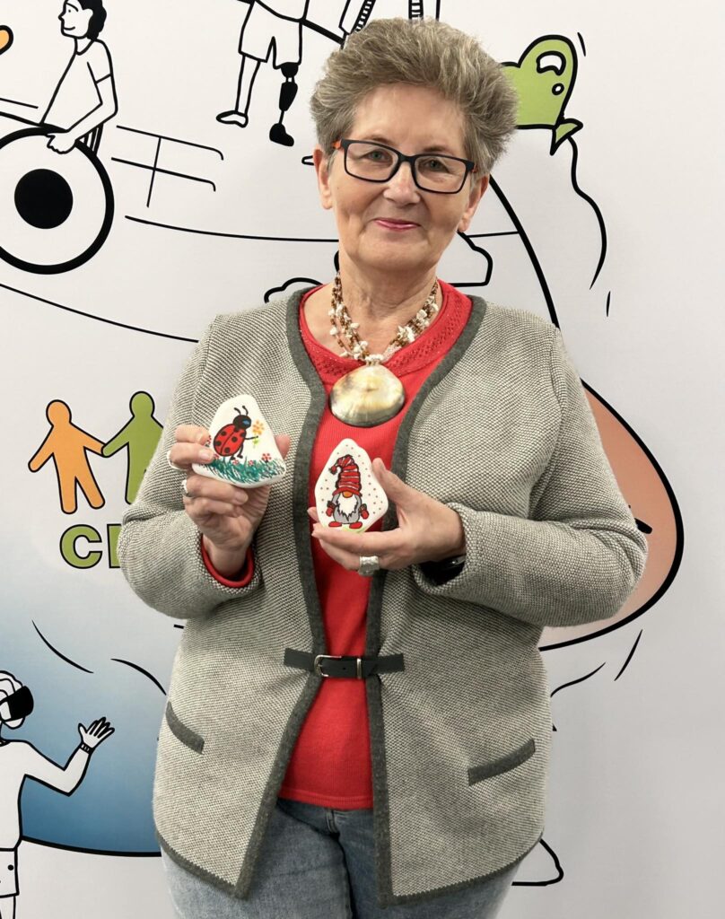 Seniorka pozuje na tle ścianki reklamowej CREO. W rękach trzyma kamyczek pomalowany farbkami.