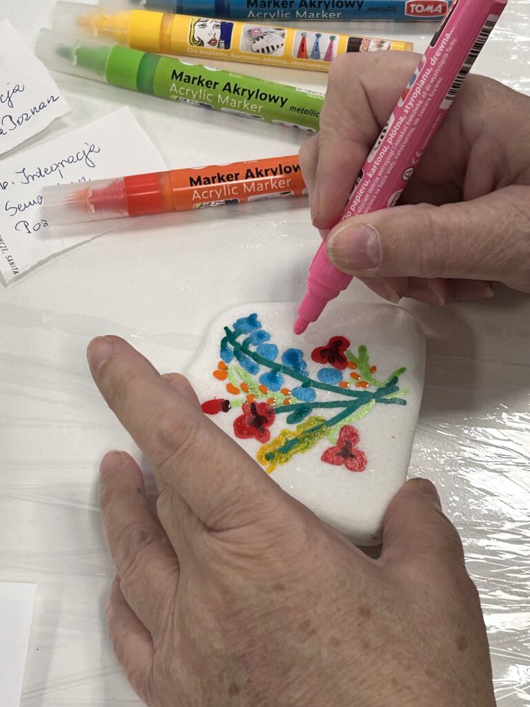 Dwie dłonie trzymają biały kamień, na którym osoba maluje kolorowe kwiaty różowowym markerem akrylowym. W tle leżą inne markery i kartki z odręcznymi notatkami.