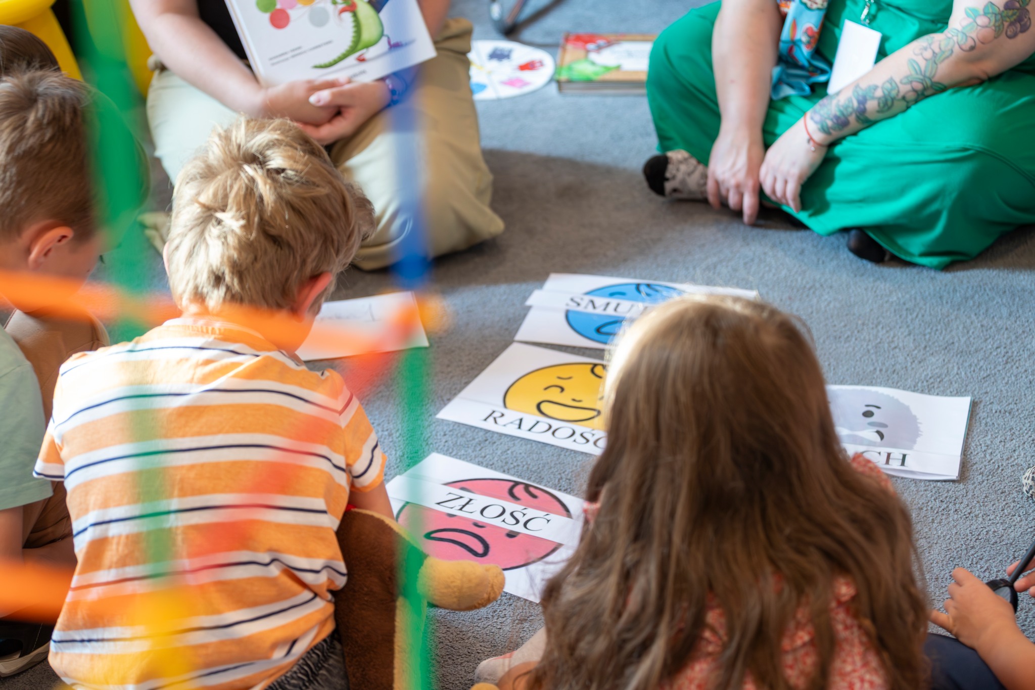 Grupa dzieci siedzi na podłodze podczas zajęć, przed sobą mają ilustracje przedstawiające różne emocje, takie jak radość, złość i smutek. Dorosłe osoby towarzyszą im, trzymając materiały edukacyjne związane z emocjami.