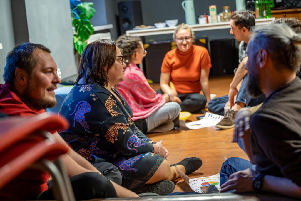 Grupa dorosłych siedzi w kręgu na podłodze podczas warsztatów, prowadząc rozmowę w swobodnej atmosferze. Uczestnicy trzymają materiały z kolorowymi grafikami, a w tle widać stół z napojami i przekąskami.