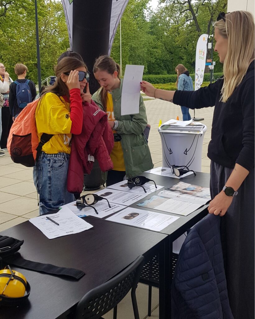 Dwoe dziewczymy stoją pod namiotem reklamowym. Przed nimi stół. Na stole symulatory wad wzroku.