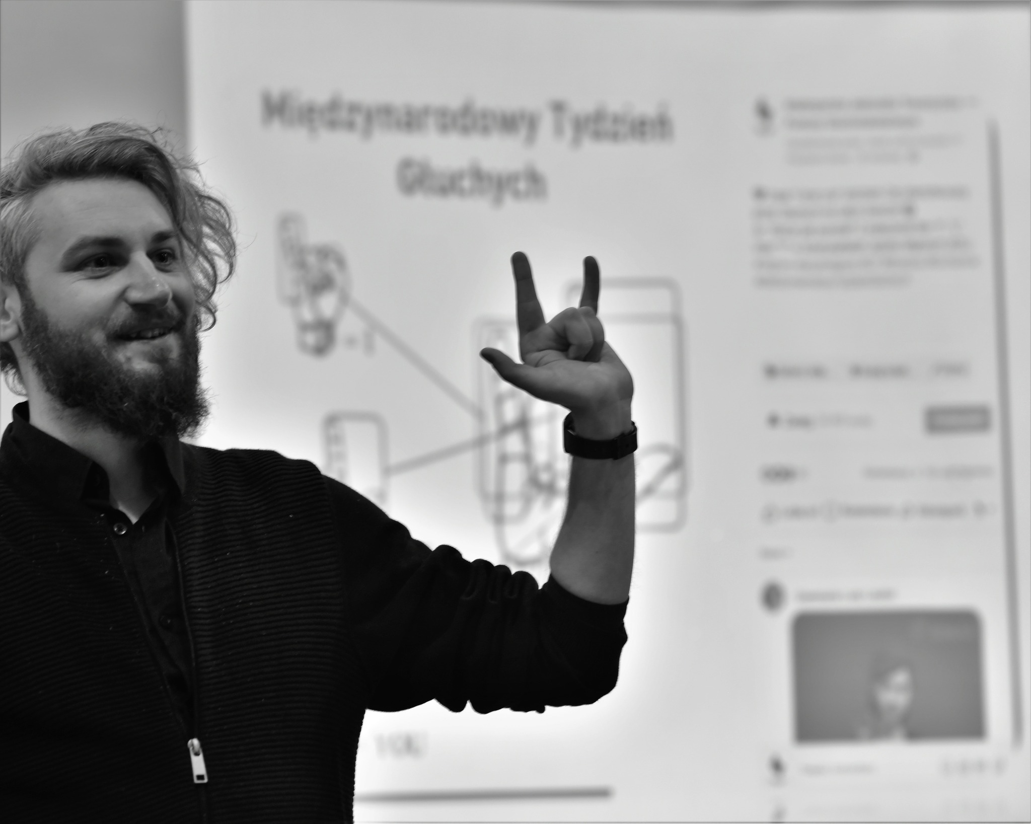 Mężczyzna pokazuje znak w języku migowym. W tle prezentacja o języku migowym